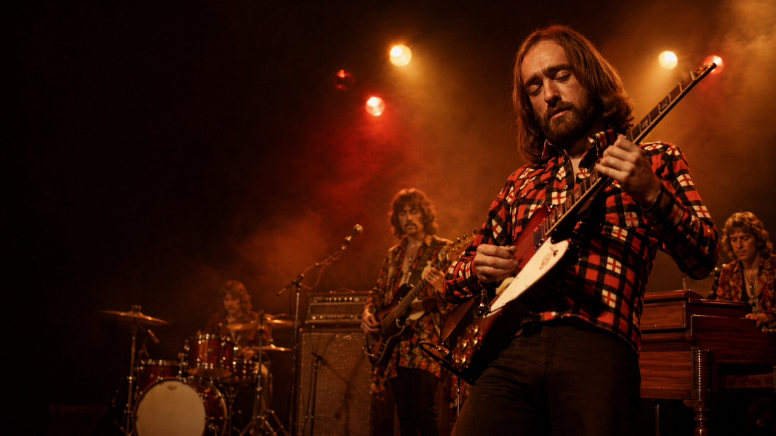 Editorial illustration of a bearded rock guitarist resembling Dave Mason performing onstage under warm red and amber lights, with drummer, bassist, and keyboard player behind him in a vintage concert setting.