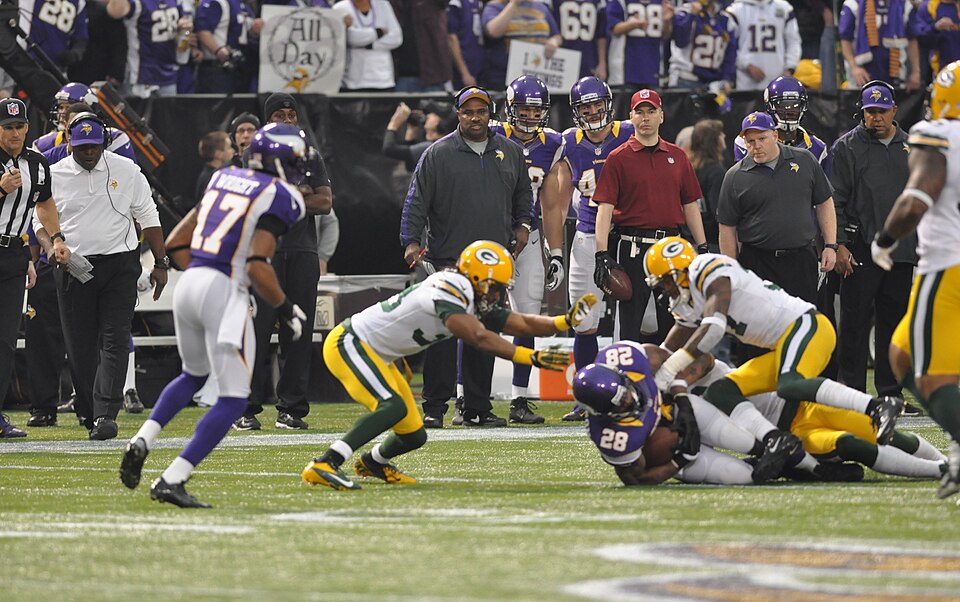 Minnesota Vikings running back Adrian Peterson is tackled by some Green Bay Packers defensive players.
On the sidelines, left to right: a referee, running backs coach James Saxon, tight ends coach Jimmie Johnson, tight ends John Carlson and Rhett Ellison, an NFL official, assistant strength and cond
