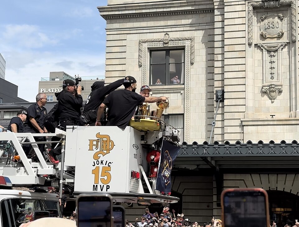 Nikola Jokić and Jamal Murphy with the Larry O'Brien Championship Trophy during the Denver Nuggets championship parade, 2023. Ladder is labeled with Jokić's name and jersey number. White building is Union Station.