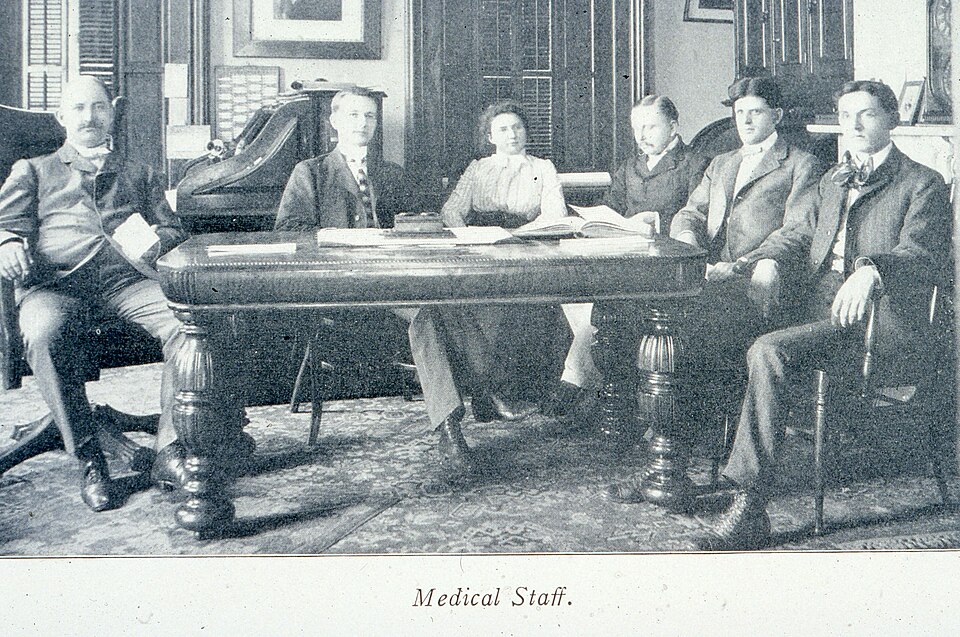 Six members of the Columbus State Hospital medical staff, including one woman, sit around an ornately carved table. A fireplace mantle can be seen to the right and a portrait hangs above a large wooden desk.