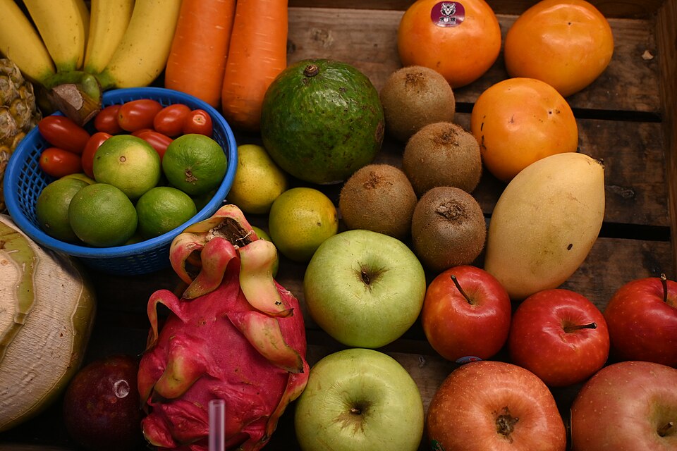 This is a vibrant display of fresh fruits and vegetables arranged on what appears to be a wooden surface. The collection includes: - Yellow bananas at the top left - Orange carrots beside them - Several oranges in the upper right - A large green avocado - Brown kiwi fruits - Various apples in green