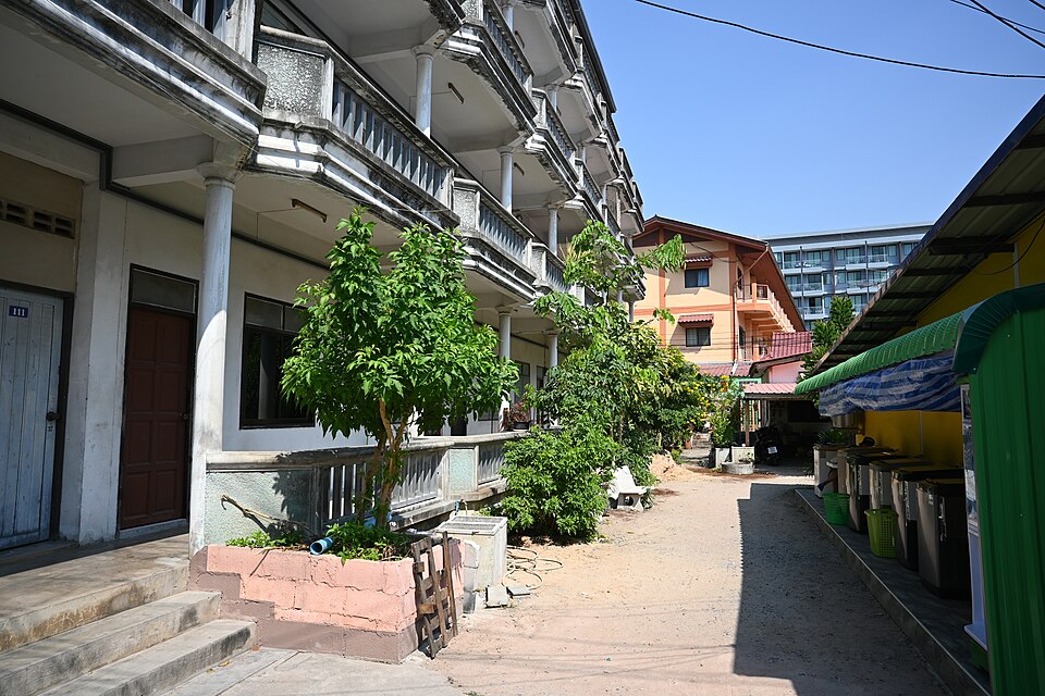 This photo shows a street view in what appears to be Pattaya City, Thailand. On the left side, there's a white multi-story building with balconies and columns, featuring a traditional Thai architectural style. Small trees with green foliage are growing in front of the building, adding some natural e