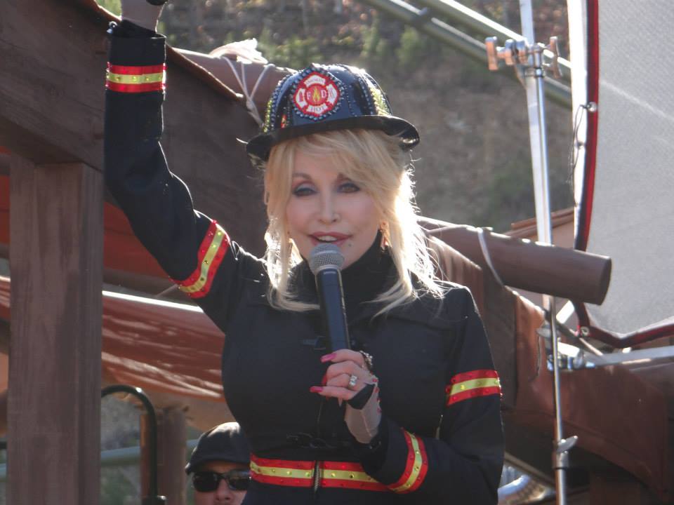 Dolly Parton, vestida con ropa de bombero, en Dollywood, presentando nuevas atracciones. Tiene un micrófono en sus manos.