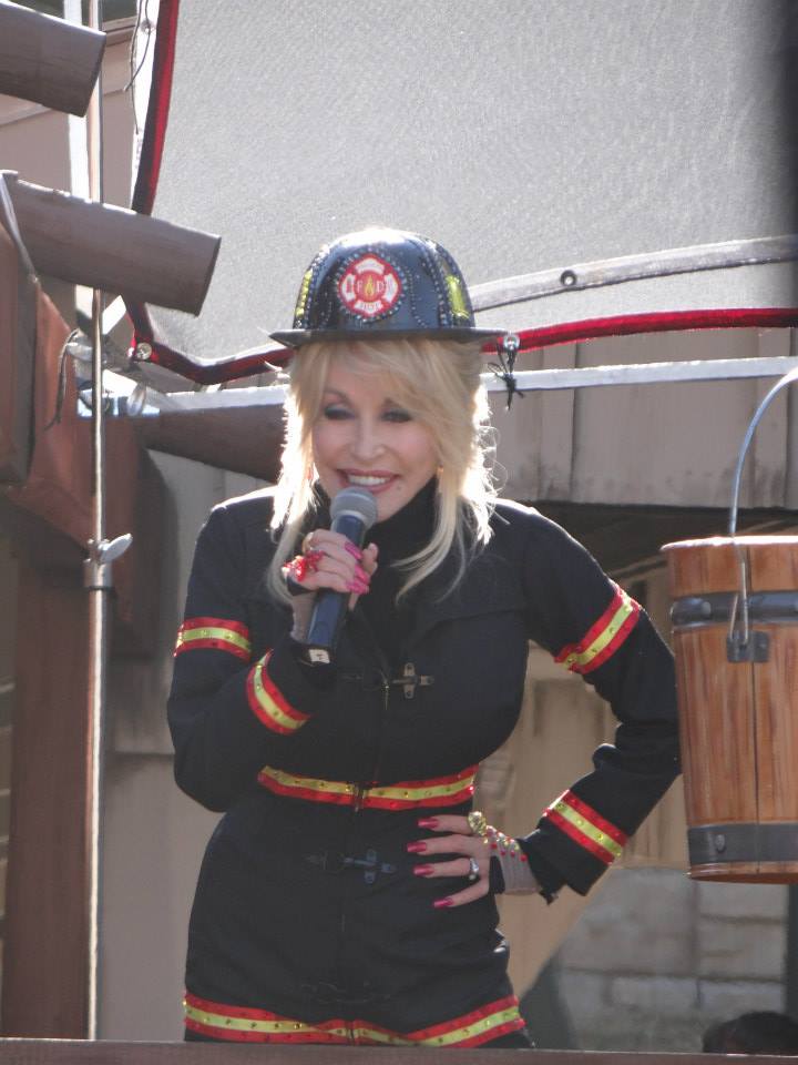 Dolly Parton, vestida con ropa de bombero, en Dollywood, presentando nuevas atracciones. Tiene un micrófono en sus manos.