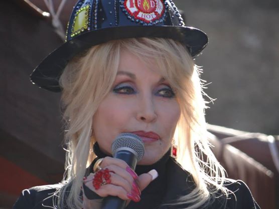Dolly Parton, vestida con ropa de bombero, en Dollywood, presentando nuevas atracciones. Tiene un micrófono en sus manos.