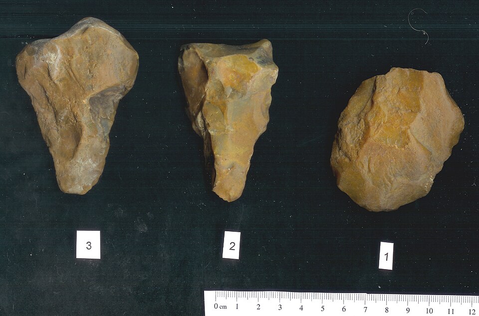 Lithic implements. 
    1. Acheulian hand axe: Flaking scars on both the ventral and dorsal sides around the edge, the tool is triangular in plan with a central point at the bottom, the butt is flat. The flaking scars are large a crude and flint has been heavily worn.  The surface patina is light br