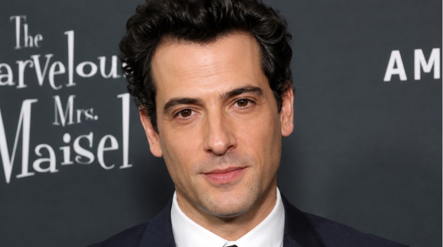 A polished red-carpet style portrait shows a Luke Kirby-inspired actor in formal attire against an entertainment event backdrop.