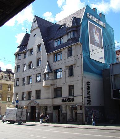 Brīvības Street, 47 (Riga)
Location: Latvia, Riga, Riga
Coordinates: 56.9566441, 24.1212638

Caption: A historic, light-colored building with ornate architecture and a dark roof stands beside a modern Samsung billboard. The "MANGO" clothing store sign is visible on the ground floor. A white delivery