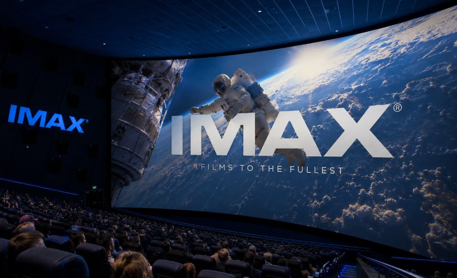 A wide interior image of an IMAX-style movie theater with rows of audience seating, blue theater lighting, and a huge curved screen showing an astronaut in orbit over Earth.