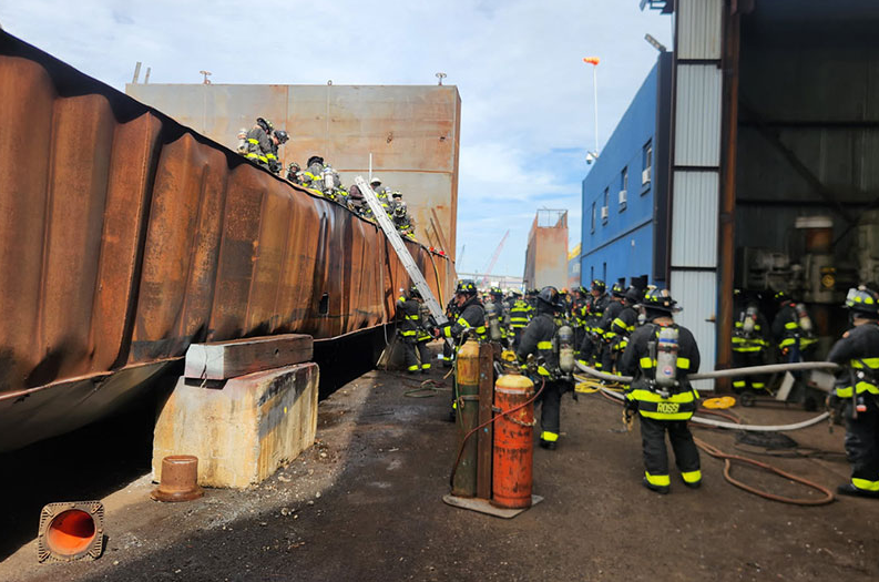 News image showing FDNY crews at the scene of a Staten Island shipyard fire and explosion that killed one person and injured more than 30 firefighters and first responders. The image shows firefighters working near a large metal structure at the Mariners Harbor site.