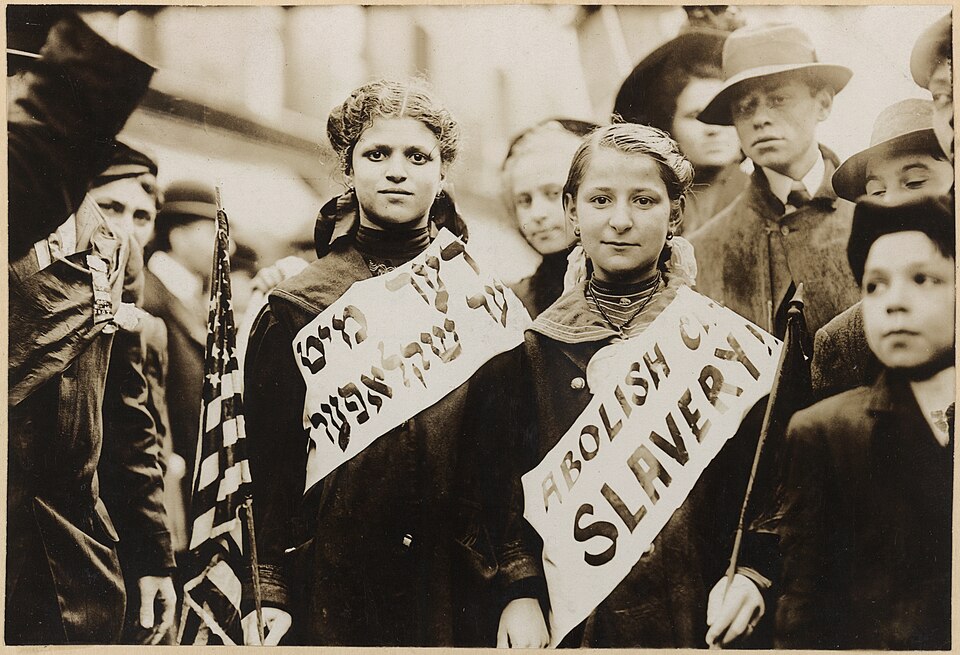 Üstünde İngilizce ve Yidiş dilinde "Çocuk Köleliğini Durdurun" sloganı yazılı olan bant taşıyan iki kız. Büyük olasılıkla 1 Mayıs 1909 tarihindeki New York işçi yürüyüşünde çekilmiş bir fotoğraf.