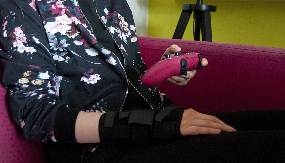 A person is sitting on a pink couch. Their body is oriented to the right along the couch. They are holding a pink PlayStation 5 controller in their left hand. On their right wrist, they are wearing a black wrist brace. The person is primarily dressed in black, and their jacket has pink and rose-colo