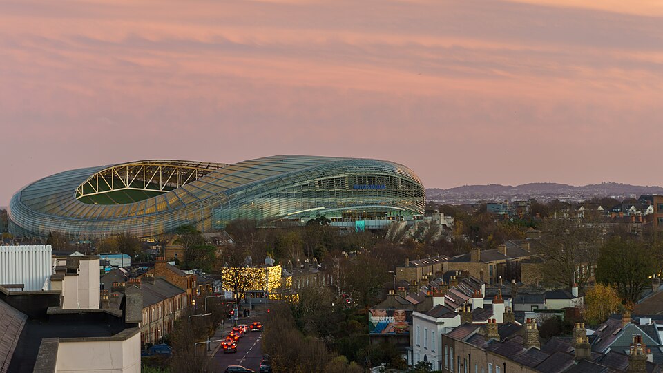 An elevated, panoramic shot capturing Aviva Stadium in Dublin, Ireland, during dusk. The stadium's distinctive glass and steel exterior reflects the soft light of a pastel pink and orange sky. In the foreground, traditional Dublin terraced houses line streets, with car headlights visible on a main r