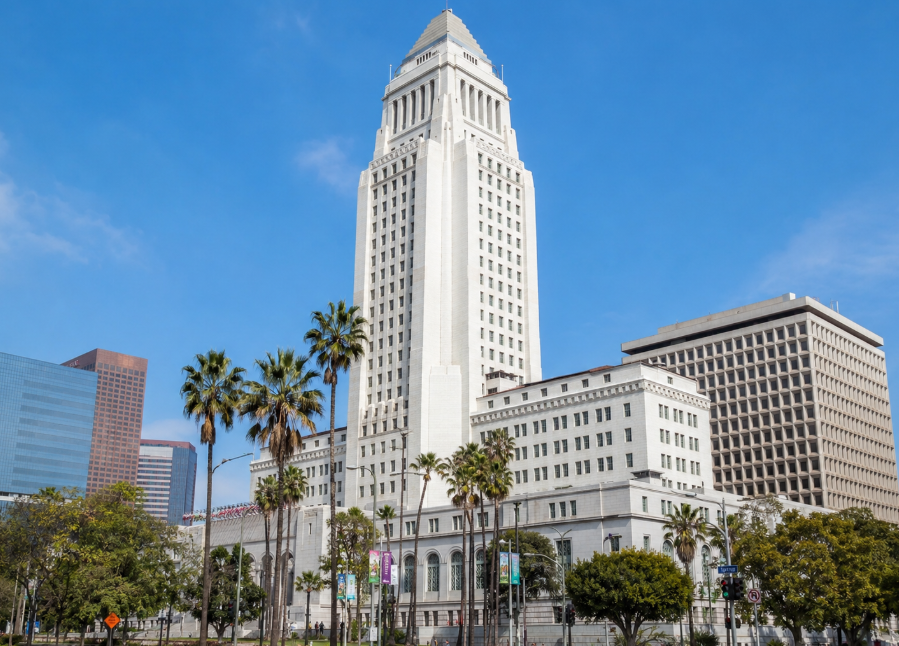 A wide daytime image of the Los Angeles City Hall building in downtown Los Angeles, showing its tall white tower, surrounding civic buildings, palm trees, and a bright blue California sky
