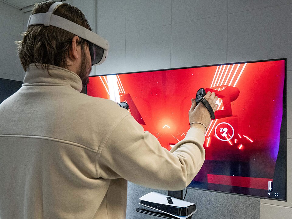 A young man is standing in front of a large screen. He is wearing a virtual reality headset and holding a controller in his right hand. The young man is positioned on the left side of the image, so only the right side of his upper body, his torso, head, and right arm are visible. He is playing the v