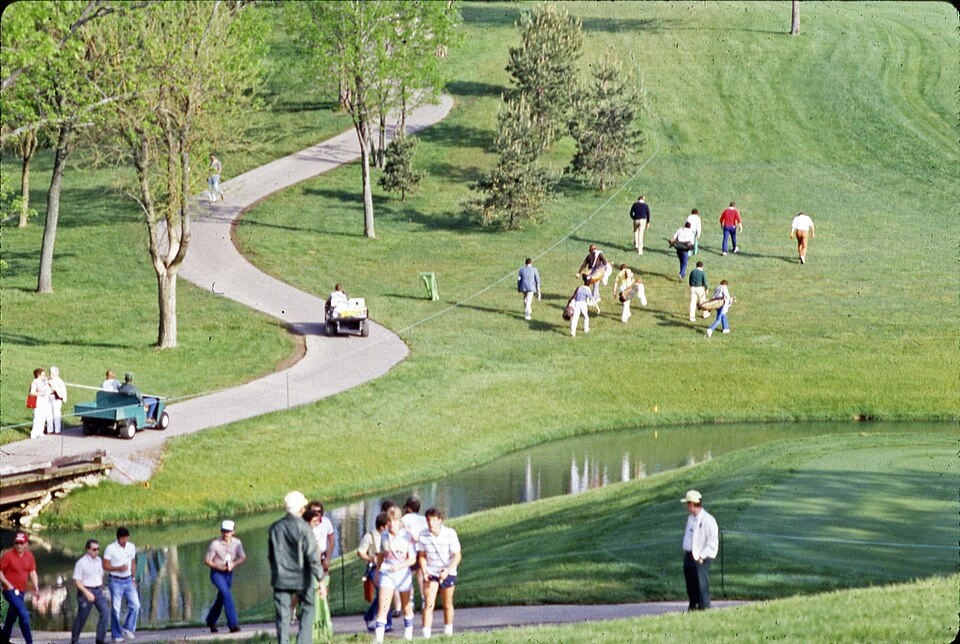 A view of an unidentified hole during the 1984 Memorial Tournament at the Muirfield Village Golf Club. This picture may have been taken during the pro-am as there looks to be five players in the group of golfers and caddie walking up the fairway. Two golf carts drive up the paved path while other sp