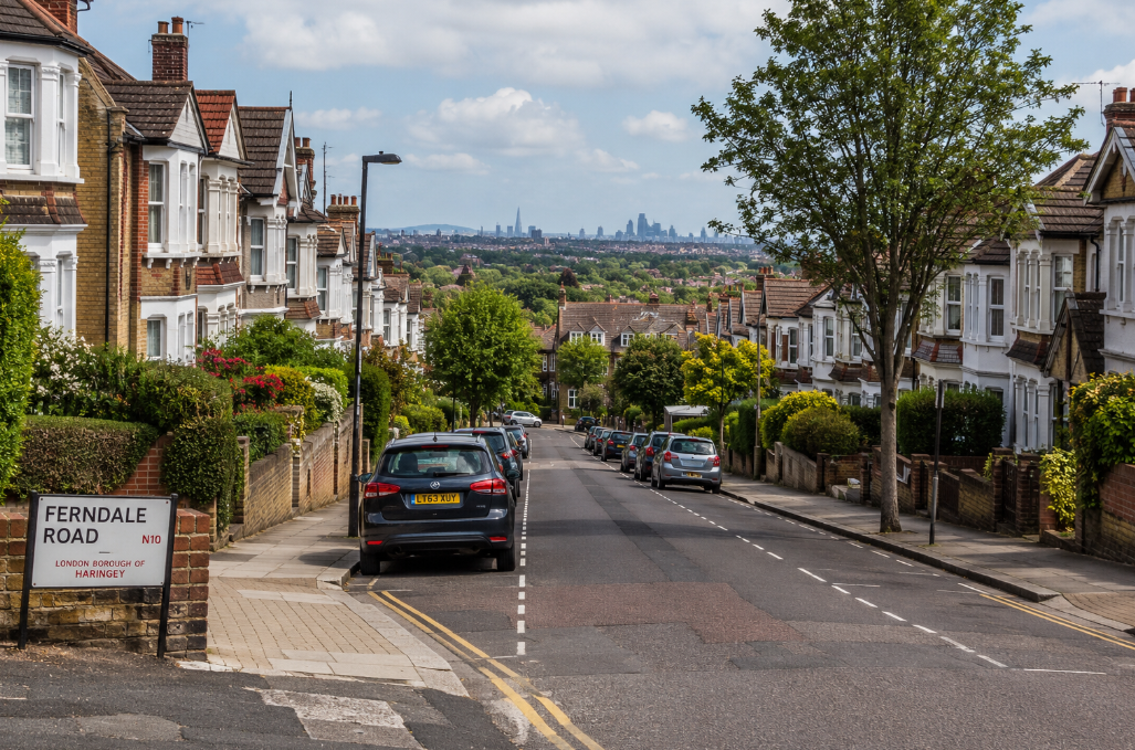 A quiet London suburban street with classic brick homes, leafy gardens, parked cars, and the distant city skyline in the background.