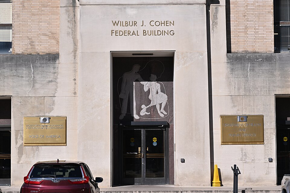 The C street entrance to the Wilbur J. Cohen building.  To the left of the door is a sign that says: "Broadcasting Board of Governors", "International Broadcasting Bureau", "Voice of America". To the right is a sign that says "Department of Health and Human Services". The U.S. Agency for Global Medi