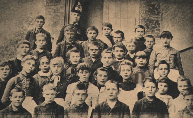 A Blade newspaper clipping dated March 24, 1957, of a class portrait of Raymer Elementary School's fifth and sixth grade students from the 1889-90 school year. The pupils from the front row, left to right, are: Frank Reichard, Charles Towers, Leroy W. Wickenden, William Corns, Thomas Blackmore, and