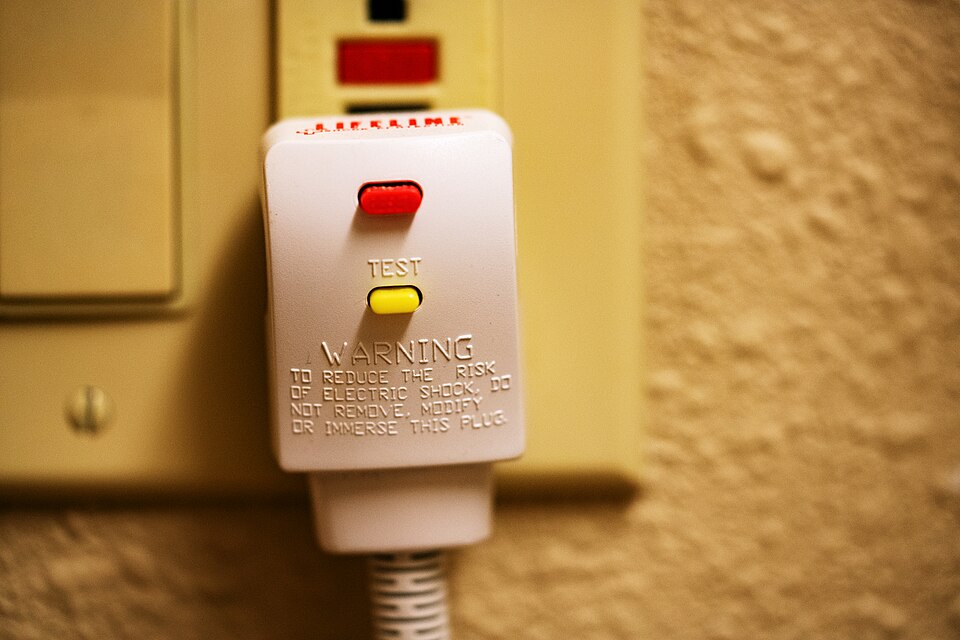 A hair dryer electric plug in an outlet, with a test and reset GFCI button. Text: test / warning / to reduce the risk of shock, do not remove, modify, or immerse this plug.