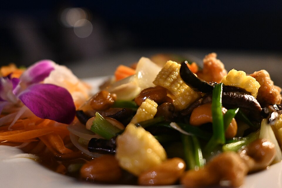 This photo shows a beautifully plated Asian stir-fry dish. The close-up view highlights a colorful mixture of ingredients including what appears to be tofu cubes, mushrooms, green vegetables (possibly green beans or scallions), carrots, and other sautéed vegetables tossed in a savory sauce. The dish