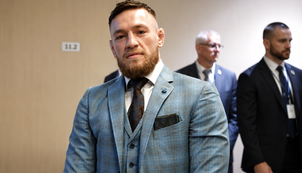 Bearded mixed martial arts fighter wearing a blue plaid three-piece suit and patterned tie in a brightly lit hallway with security personnel behind him.