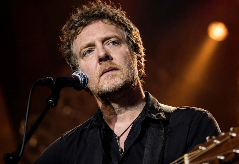 Irish musician Glen Hansard performs onstage with an acoustic guitar and microphone under warm concert lighting.