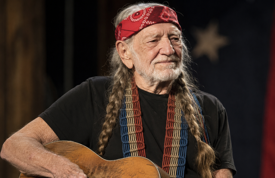 Willie Nelson wearing a red bandana and holding his well-worn acoustic guitar against a softly lit Americana background.
