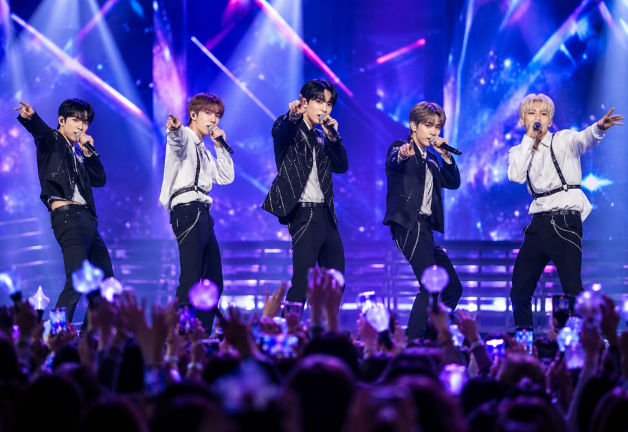 A generic K-pop band performs on a brightly lit stage in front of a cheering audience holding light sticks.