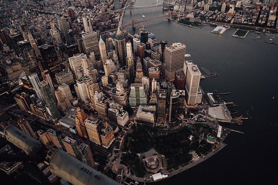 High-angle aerial view of the Financial District in Lower Manhattan, New York City, taken at dusk. The image shows the urban density of the skyscrapers along Water Street and State Street, leading down to Battery Park at the southern tip of Manhattan. The Whitehall Terminal (Staten Island Ferry) is