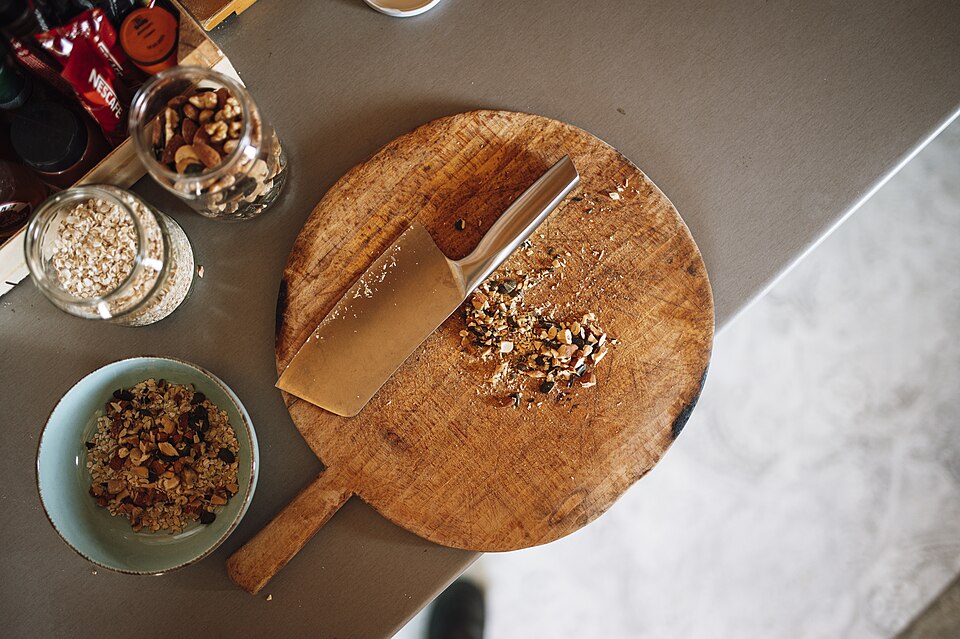 A wooden board is placed on a table with a kitchen knife on it. Nuts and herbs are chopped and scattered. Nearby, there are jars and a bowl with mixed ingredients.