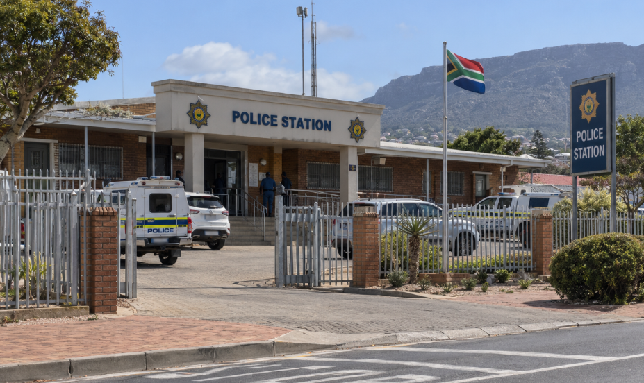 Exterior of a South African police station with police vehicles, security fencing, and the South African flag flying outside.